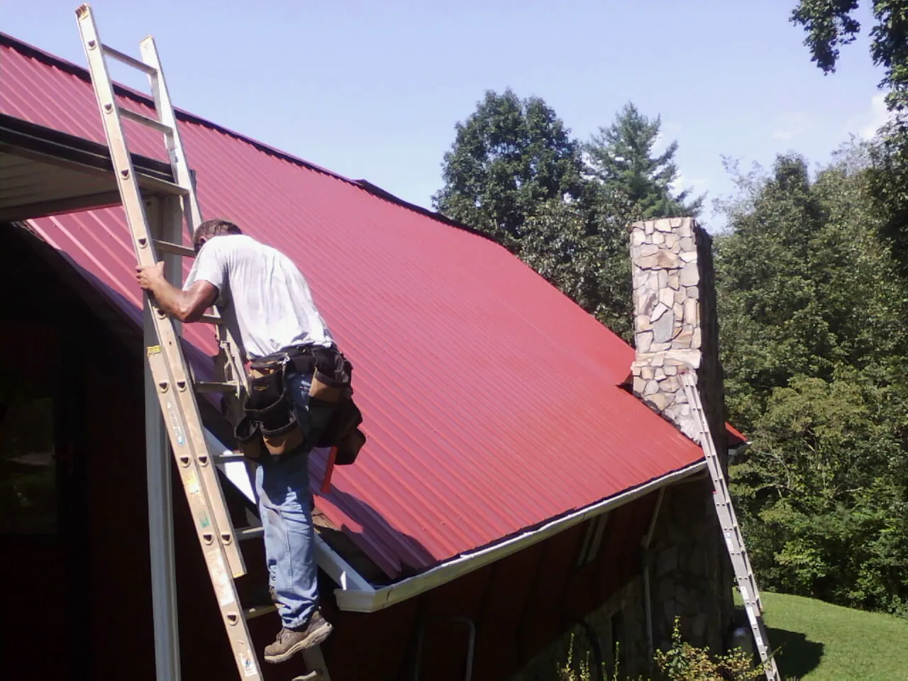 Roofer on a ladder beside a red metal roof and stone chimney — Soffit & Fascia Repair by Roofing Brothers in Oak Ridge